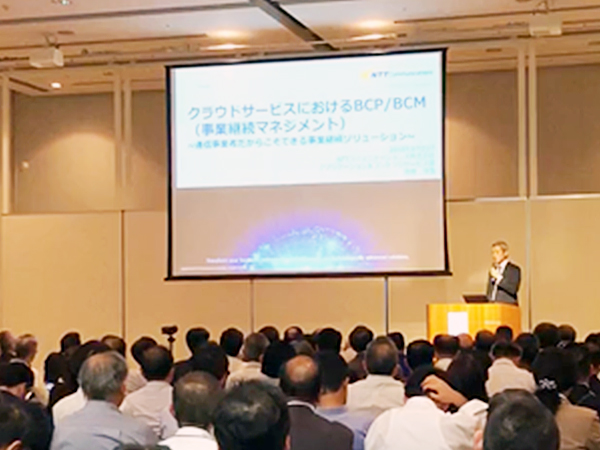 資料が映し出されている大きなスクリーンと登壇者に向かって大勢の人が座りながら視聴している様子